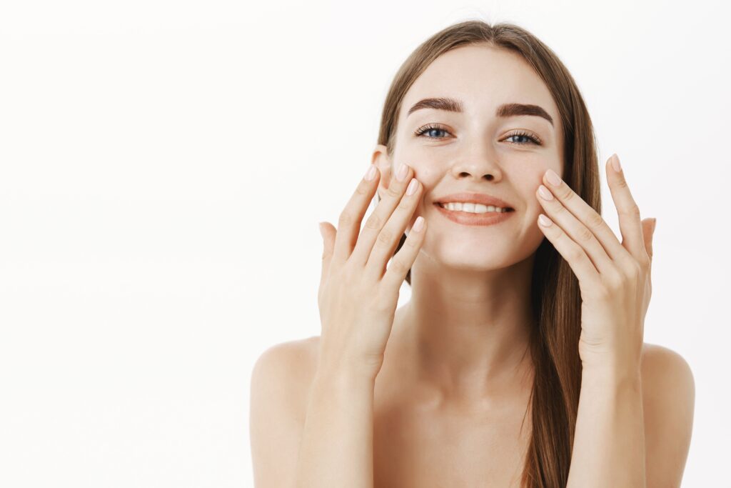 charming-relaxed-and-gentle-young-woman-making-cosmetological-procedure-applying-facial-cream-on-face-with-fingers-and-smiling-broadly-feeling-perfect-taking-care-of-skin (1) (1)